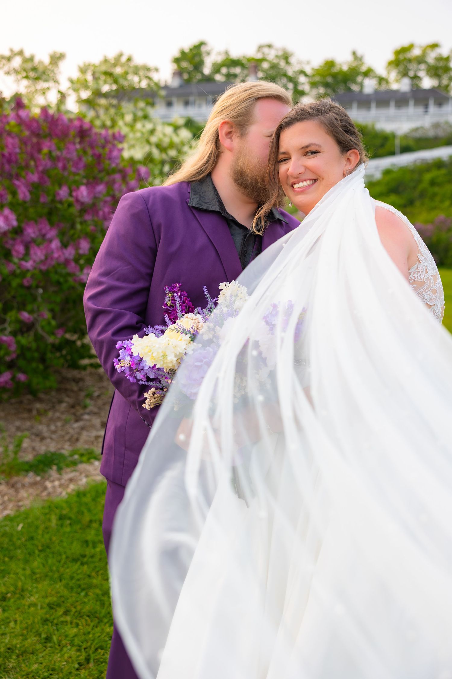 A couple in wedding attire share intimate moments in a garden setting with purple flowers in the background.