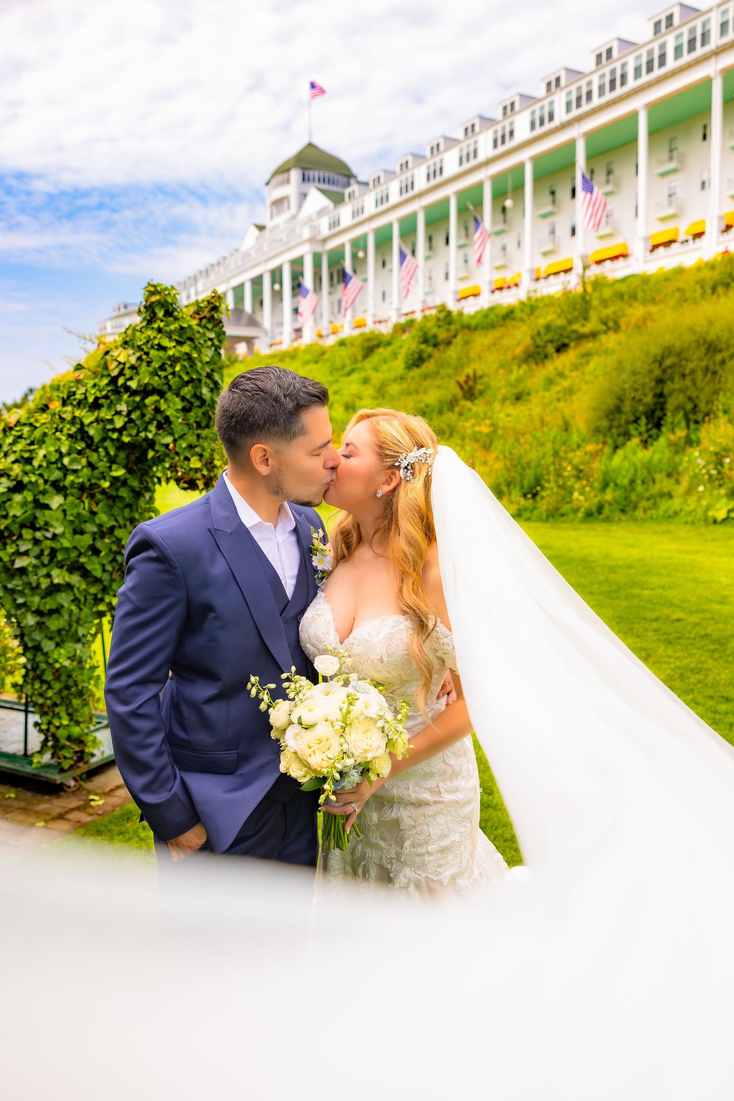A romantic wedding couple shares a kiss on a grassy hillside with a grand white hotel building in the background.