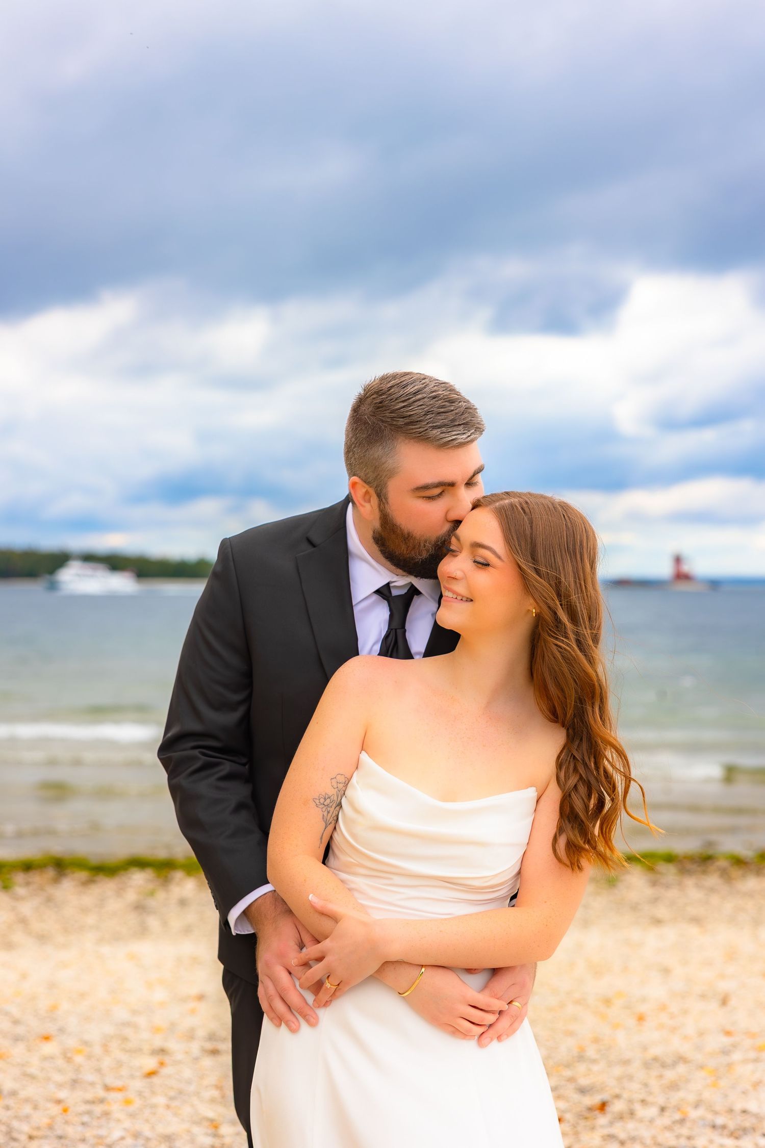 A romantic couple in wedding attire share intimate moments on a beach with cloudy skies and ocean in the background.