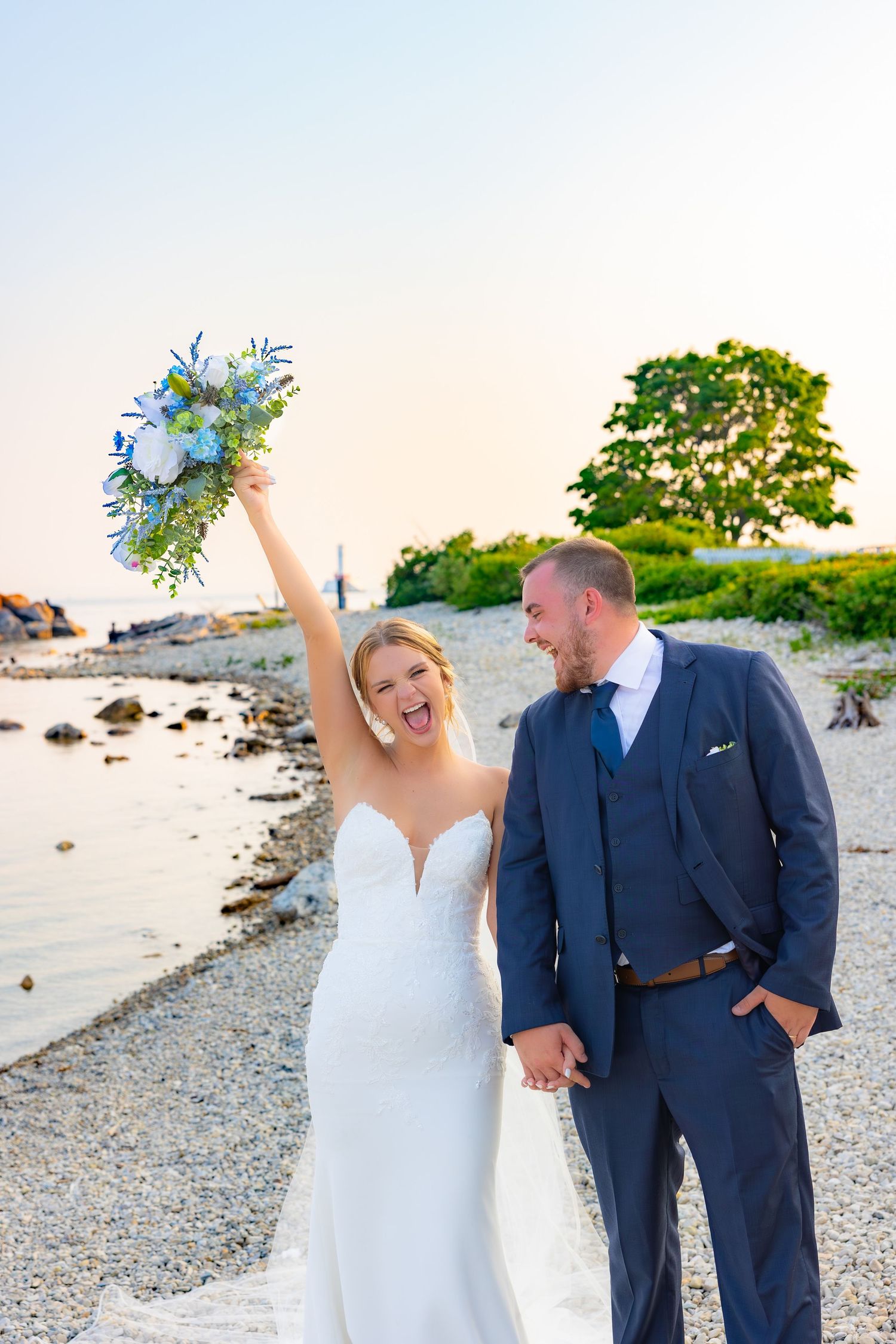 A couple celebrates joyfully on a rocky beach at sunset wearing formal wedding attire in navy and white.