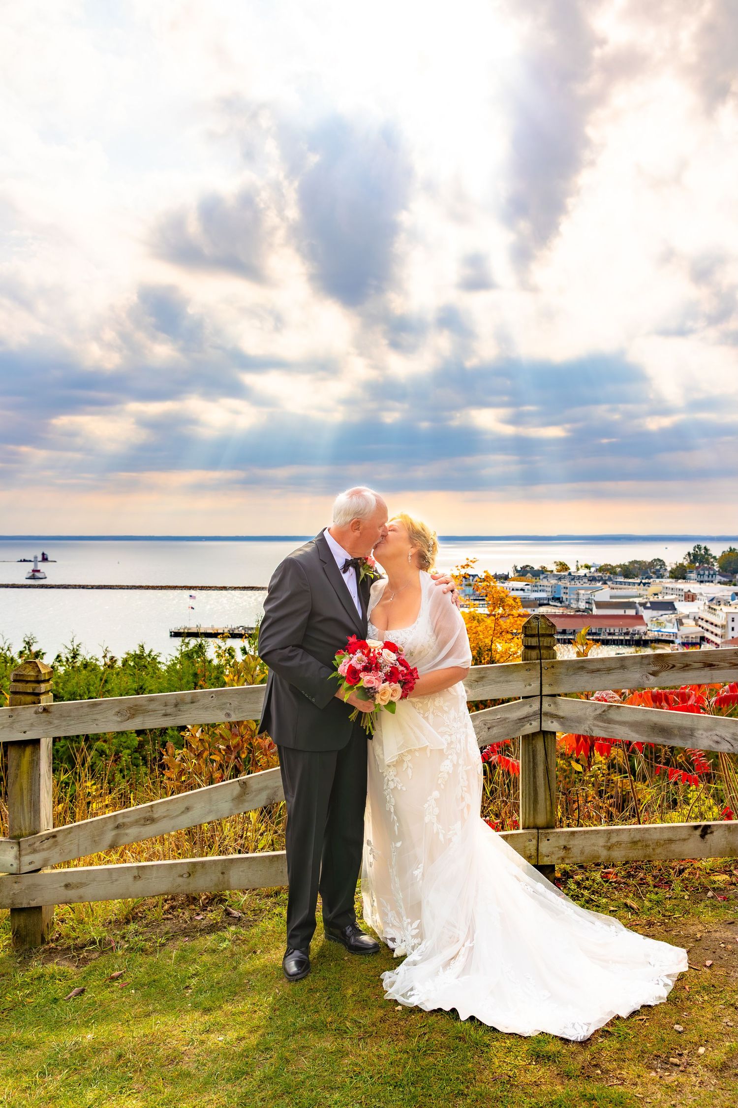 A romantic wedding couple embraces on a scenic coastal boardwalk with autumn flowers and water views at sunset.
