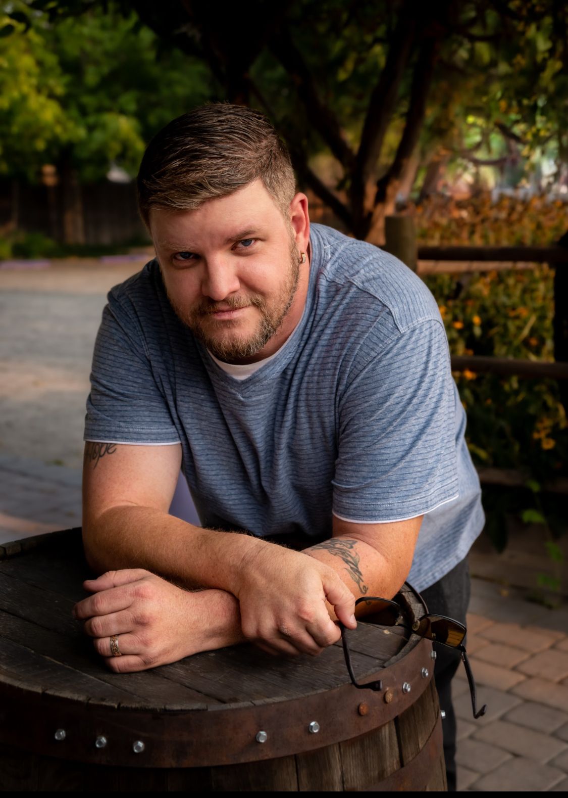 A smiling man with a beard leans on a wooden barrel outdoors, wearing a blue t-shirt with a tattoo visible on his arm.