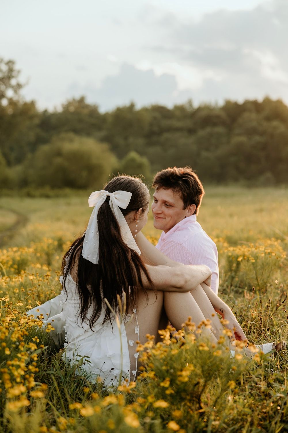 A romantic couple sits together in a wildflower field during golden hour as the sun sets behind them.