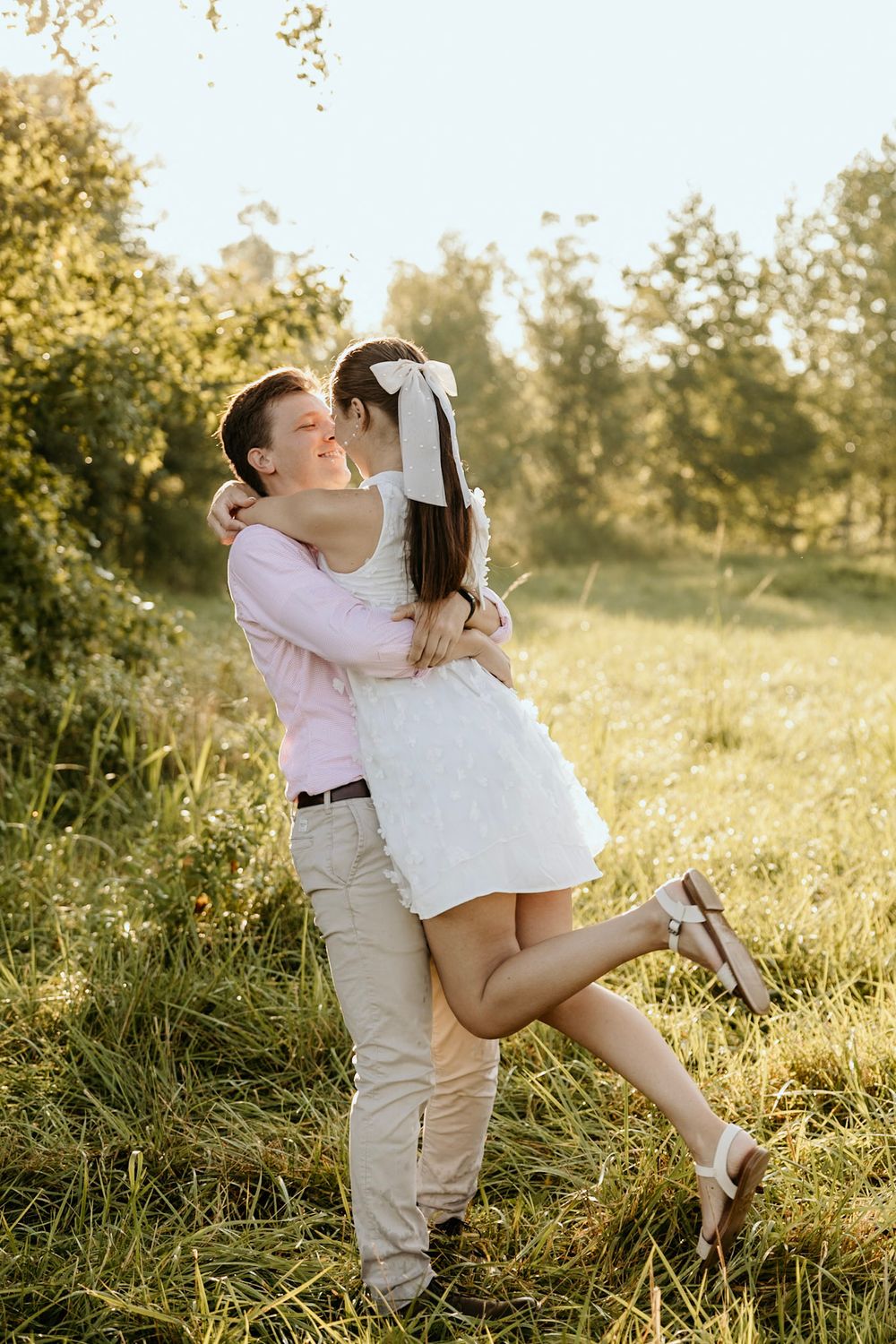 Young couple embraces romantically in a sunlit meadow during golden hour, with the woman wearing a white dress.