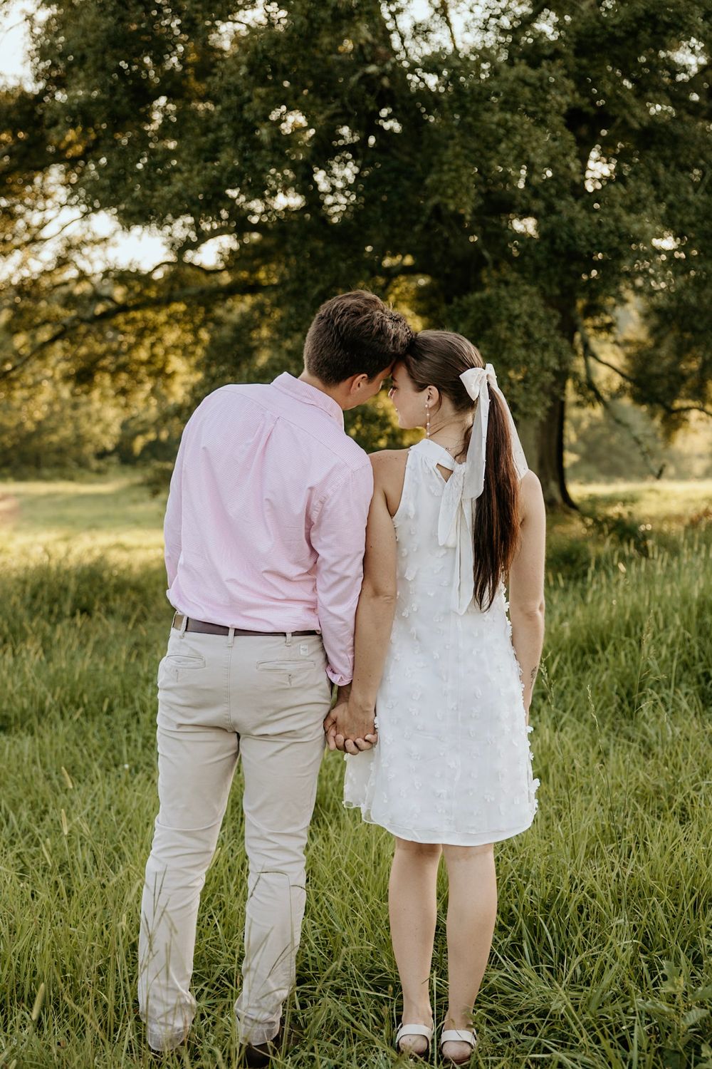 Couple holding hands in a grassy field at sunset with trees in the background.