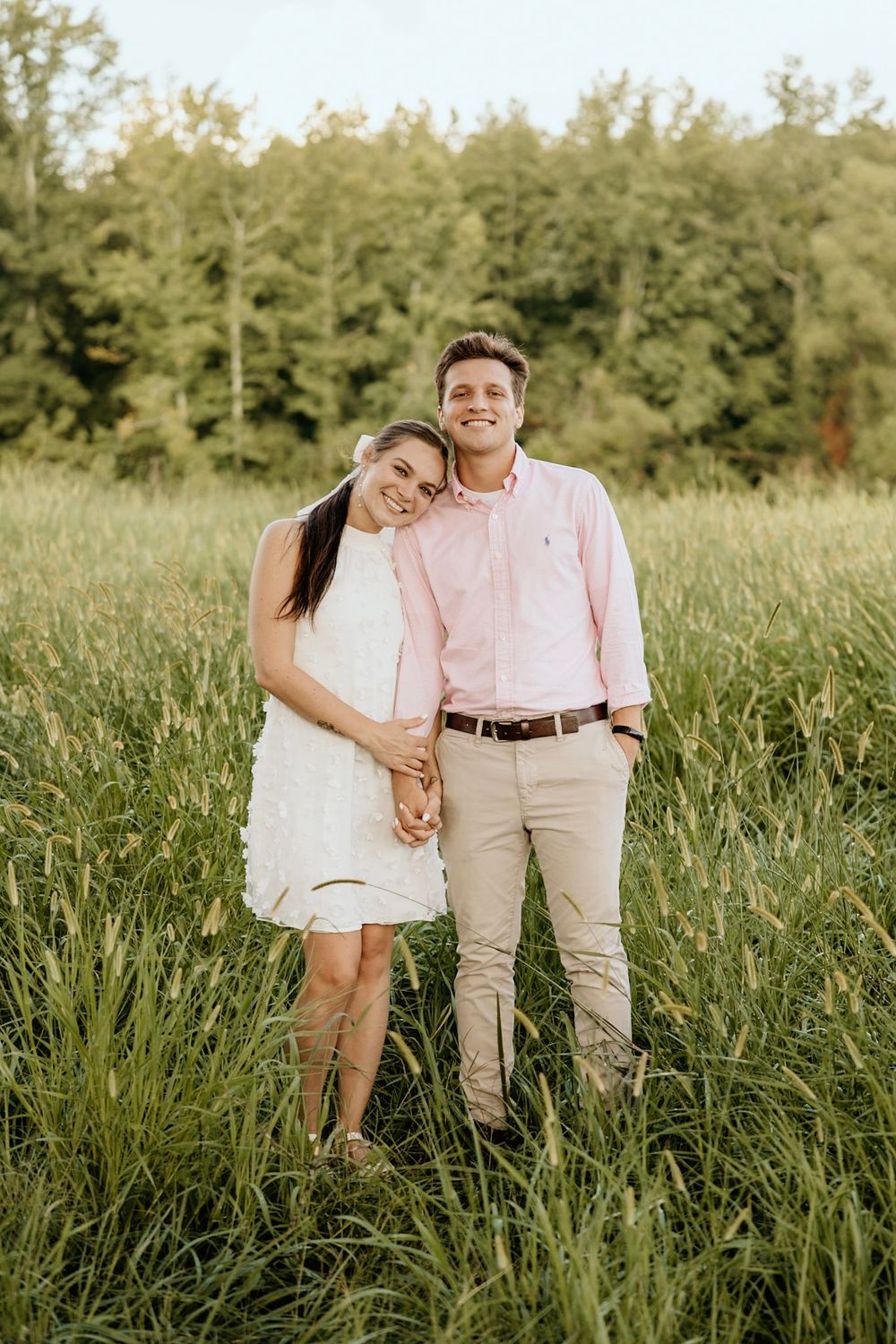 A couple wearing coordinating pink and white outfits stands together in a sun-lit meadow with trees in the background.