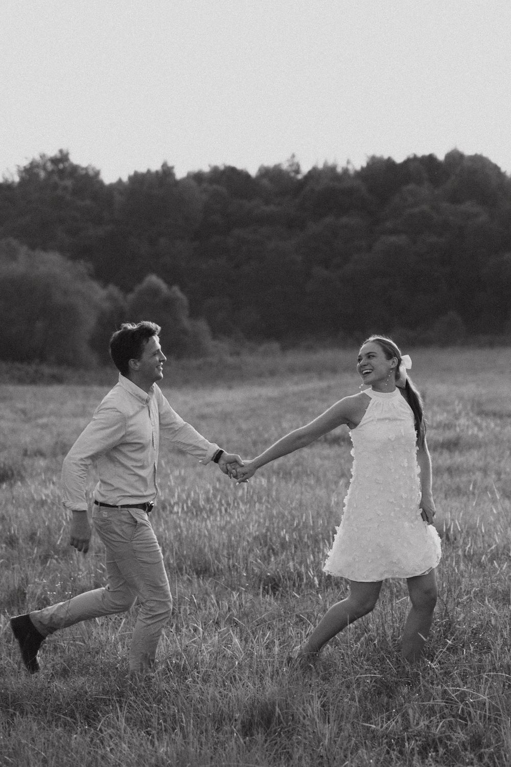 A couple in black and white holds hands while running through a grassy meadow at dusk with trees in background.