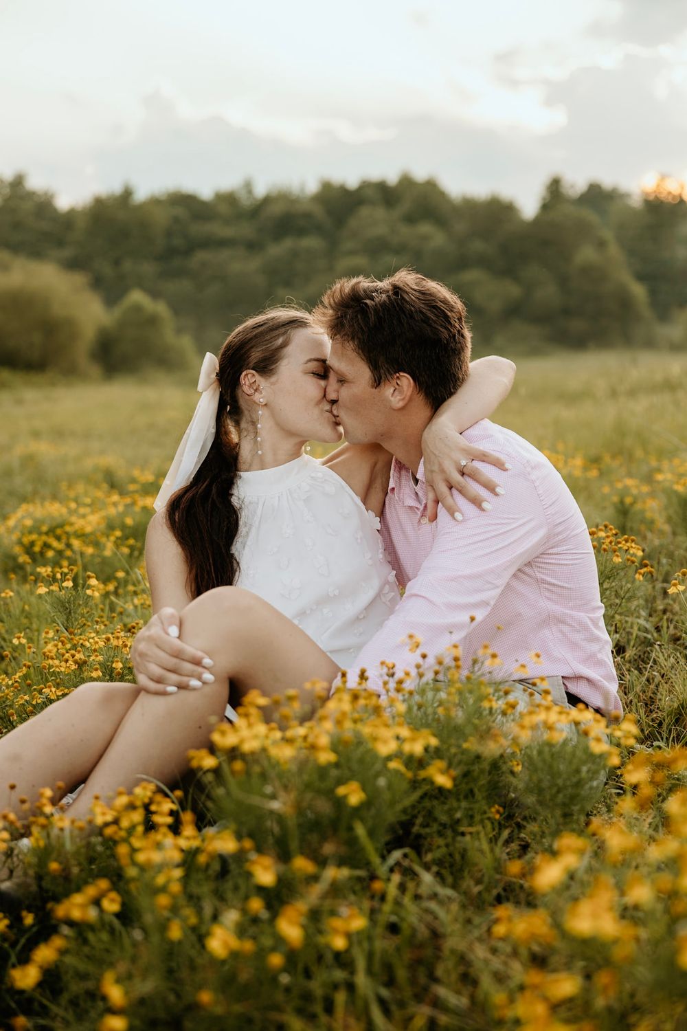 A couple shares a romantic kiss while sitting in a beautiful meadow filled with blooming yellow wildflowers at sunset.