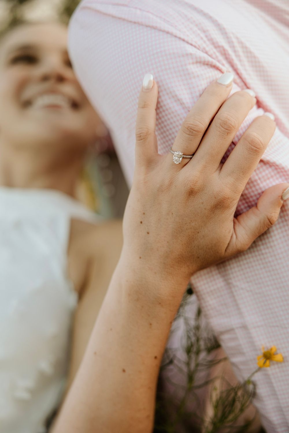 A delicate hand wearing a simple wedding band rests against light fabric with a small yellow flower in the background.
