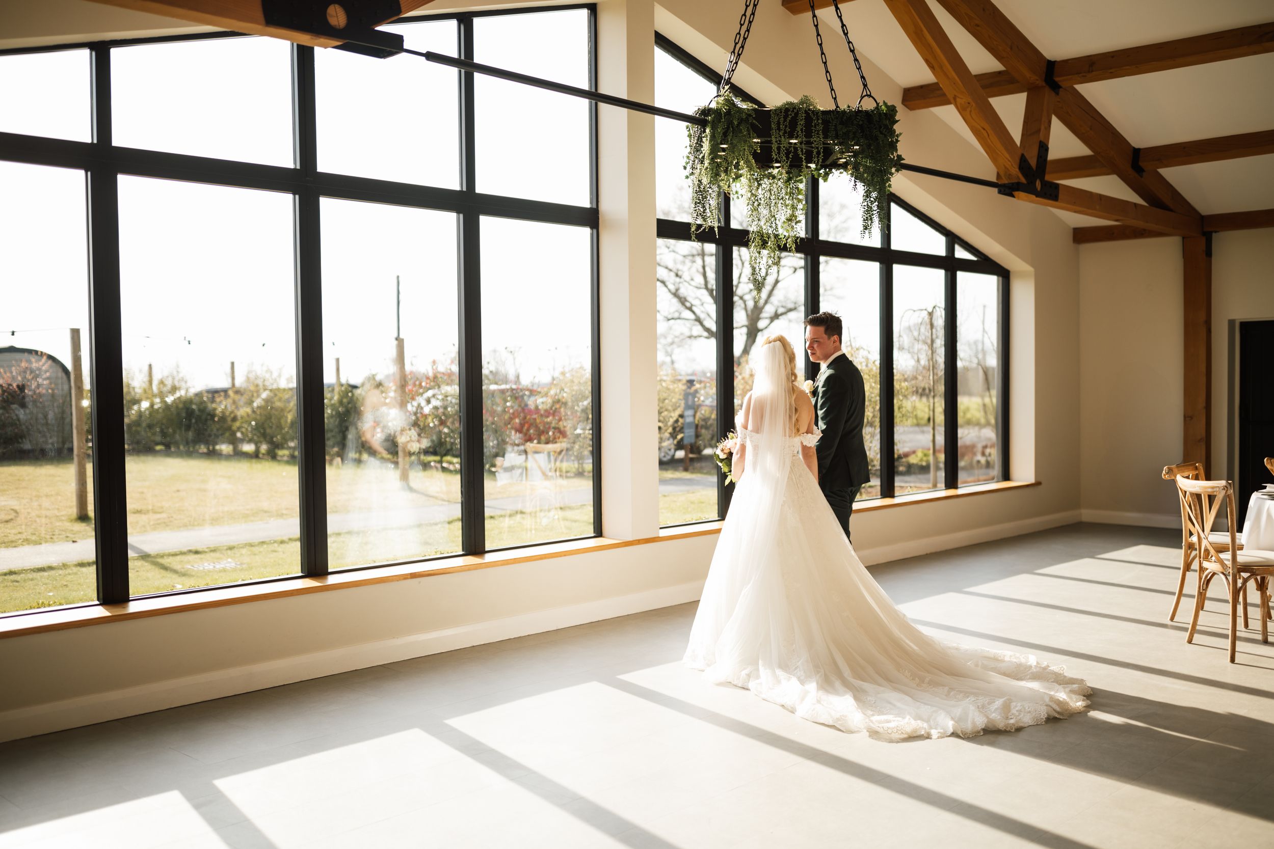 A bride in a flowing white gown stands silhouetted against large windows in a rustic venue with wooden beams.