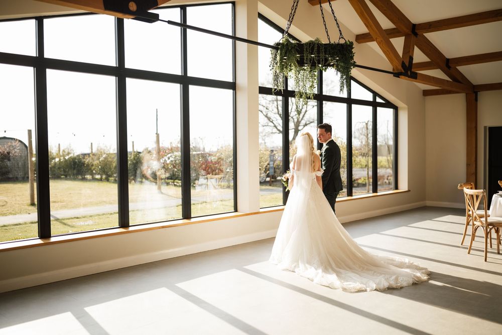 A bride in a flowing white gown stands silhouetted against large windows in a rustic venue with wooden beams.