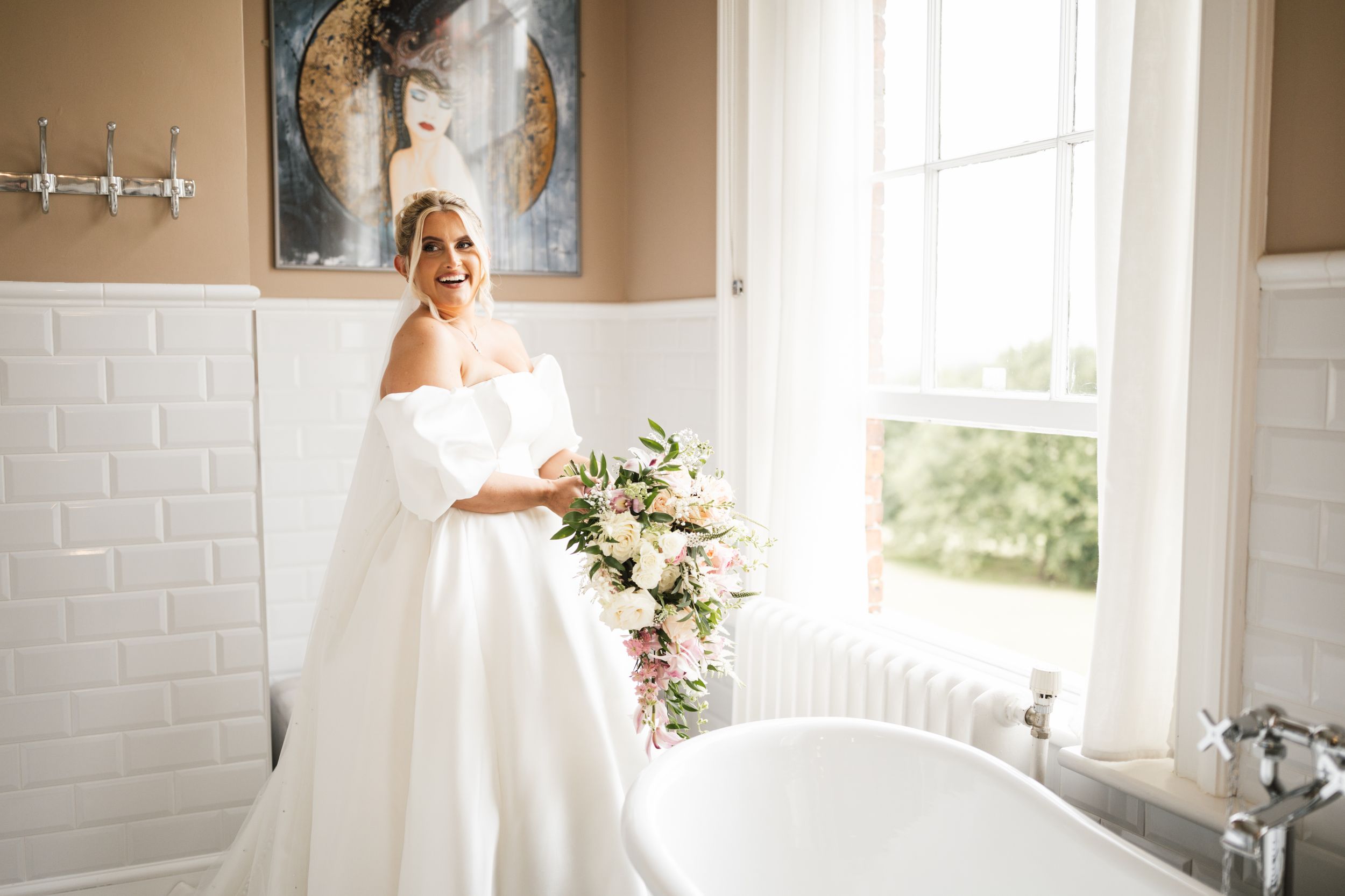 A bride in a flowing white wedding dress stands near a bright window in a classic white-tiled room.