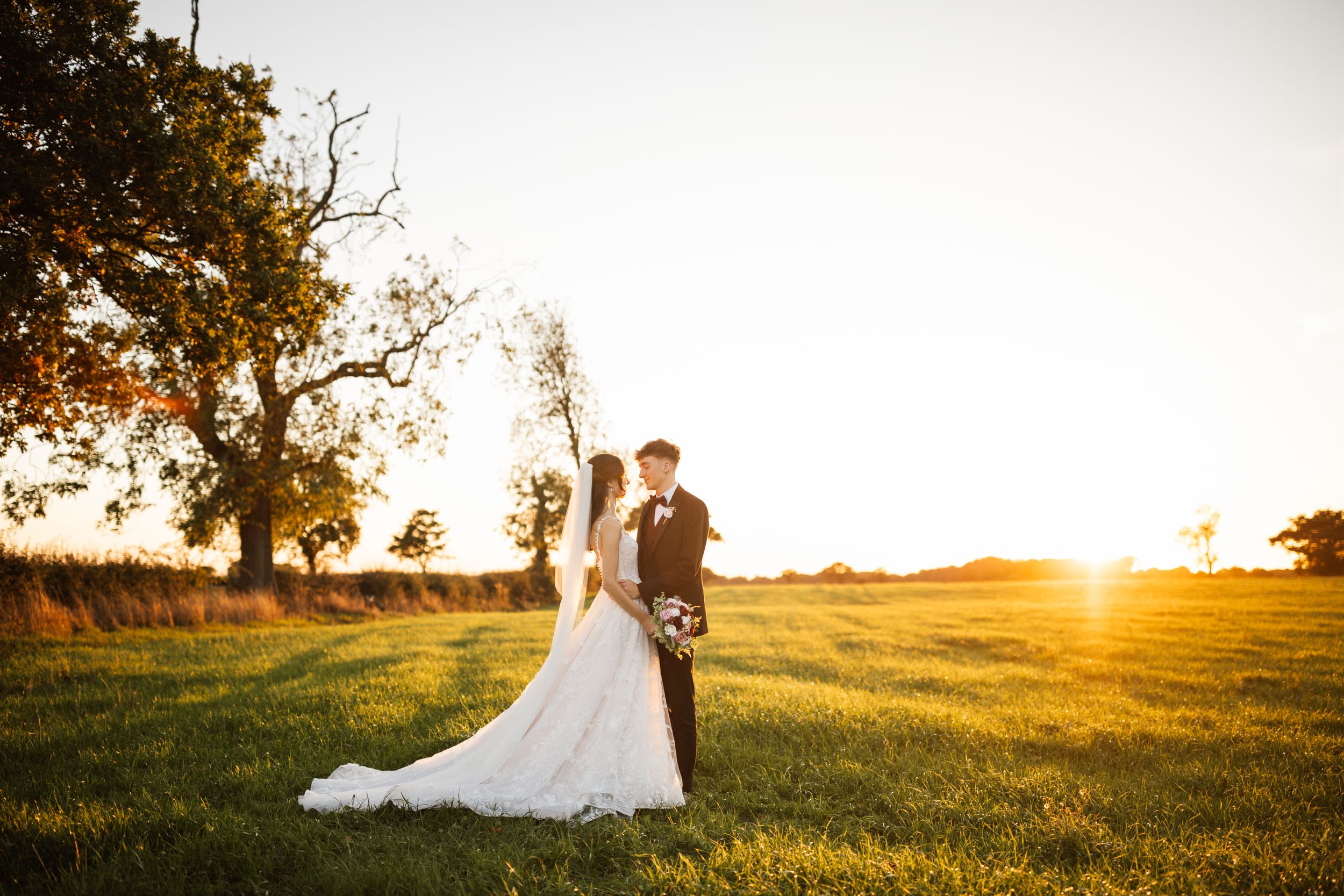 A romantic couple stands in a golden sunlit field during sunset while a white wedding dress flows in the breeze.