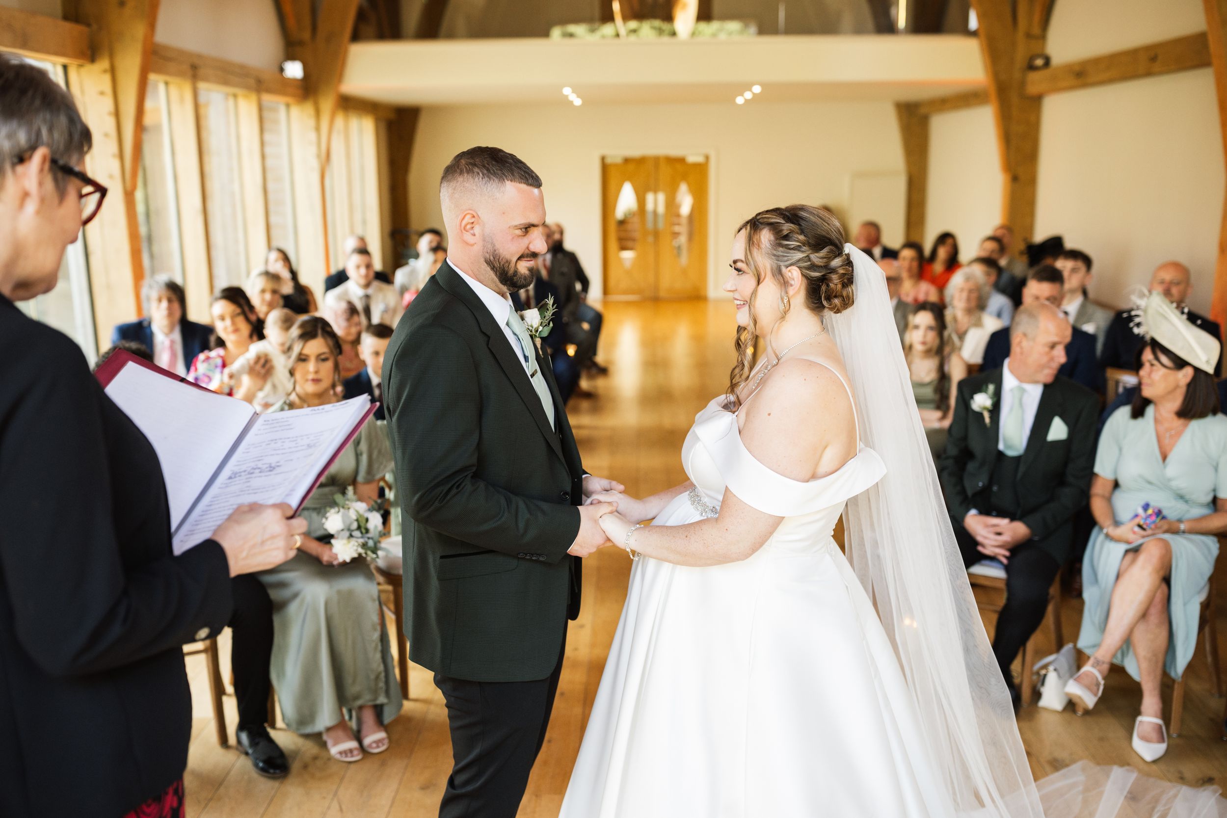 A joyful couple exchanges wedding vows during an intimate ceremony in a wooden beam venue with natural light.