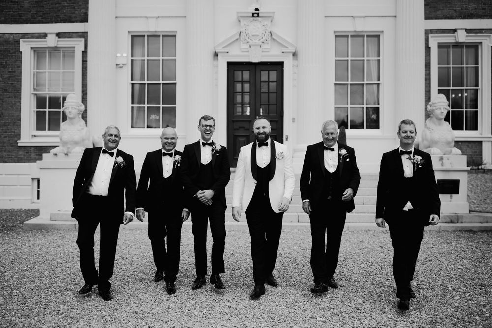 Groomsmen in black tuxedos walk across a white building facade in a candid wedding photo in black and white.