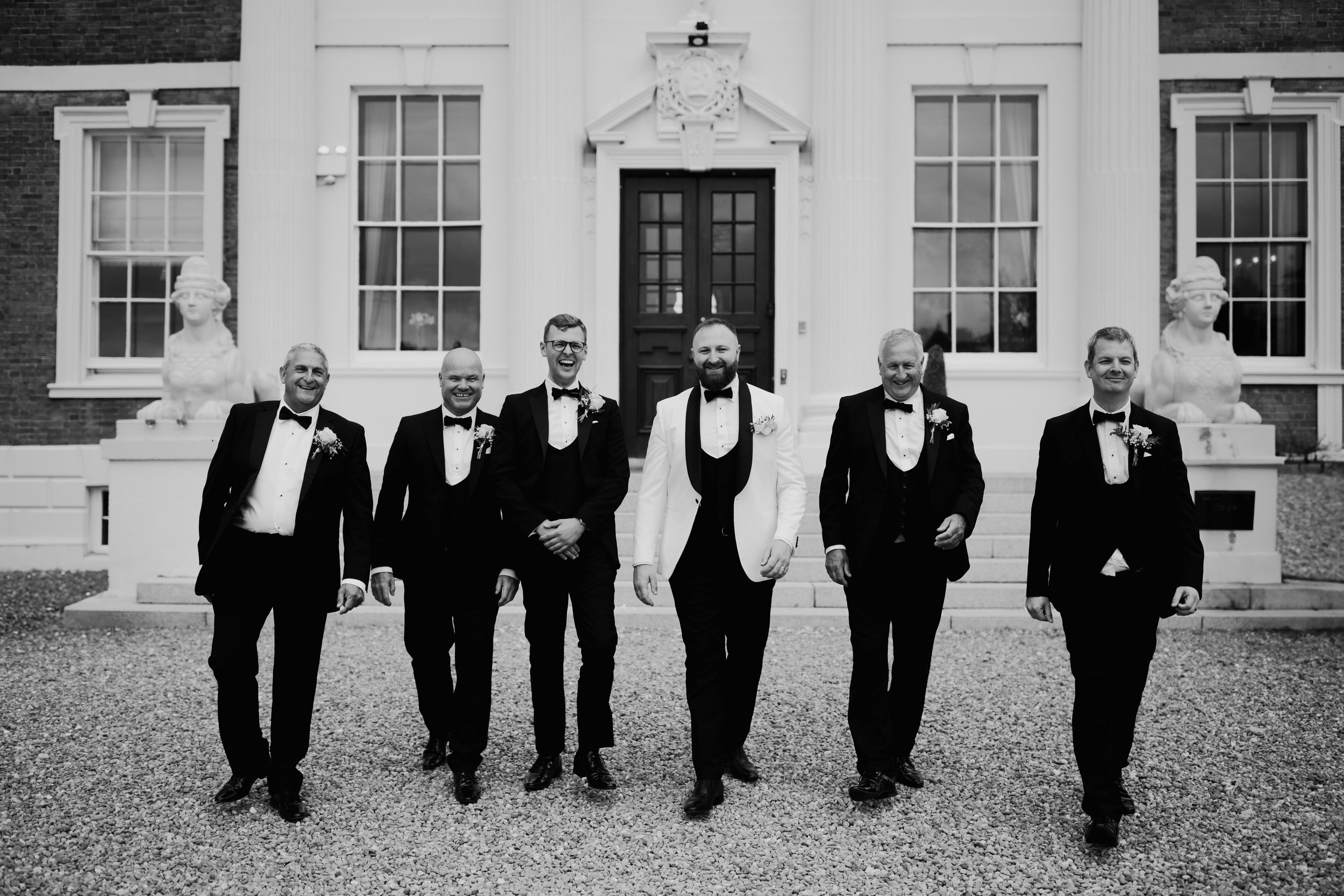 Groomsmen in black tuxedos walk across a white building facade in a candid wedding photo in black and white.