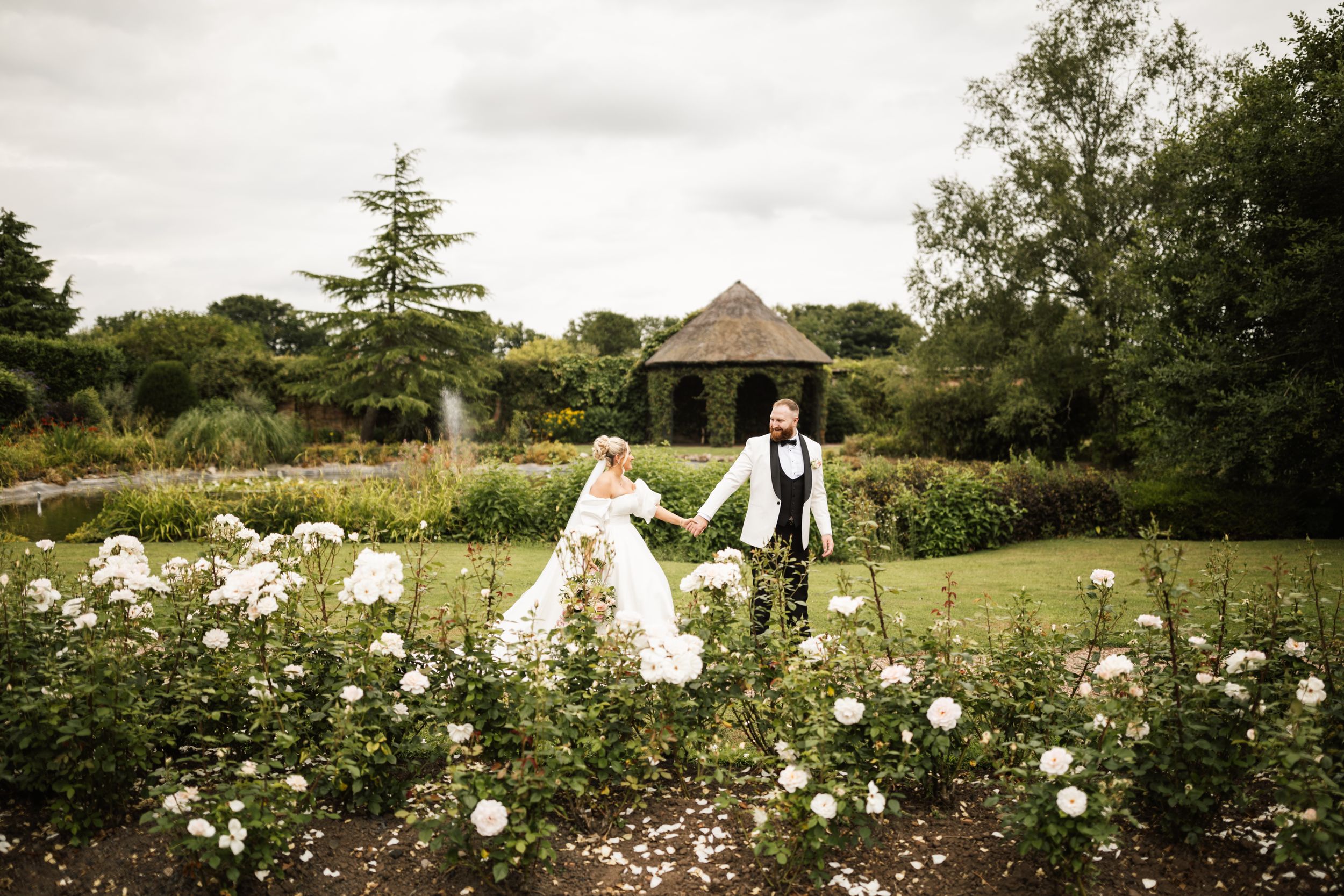 A couple walks through a lush garden with white roses and a gazebo in the background on an overcast day.