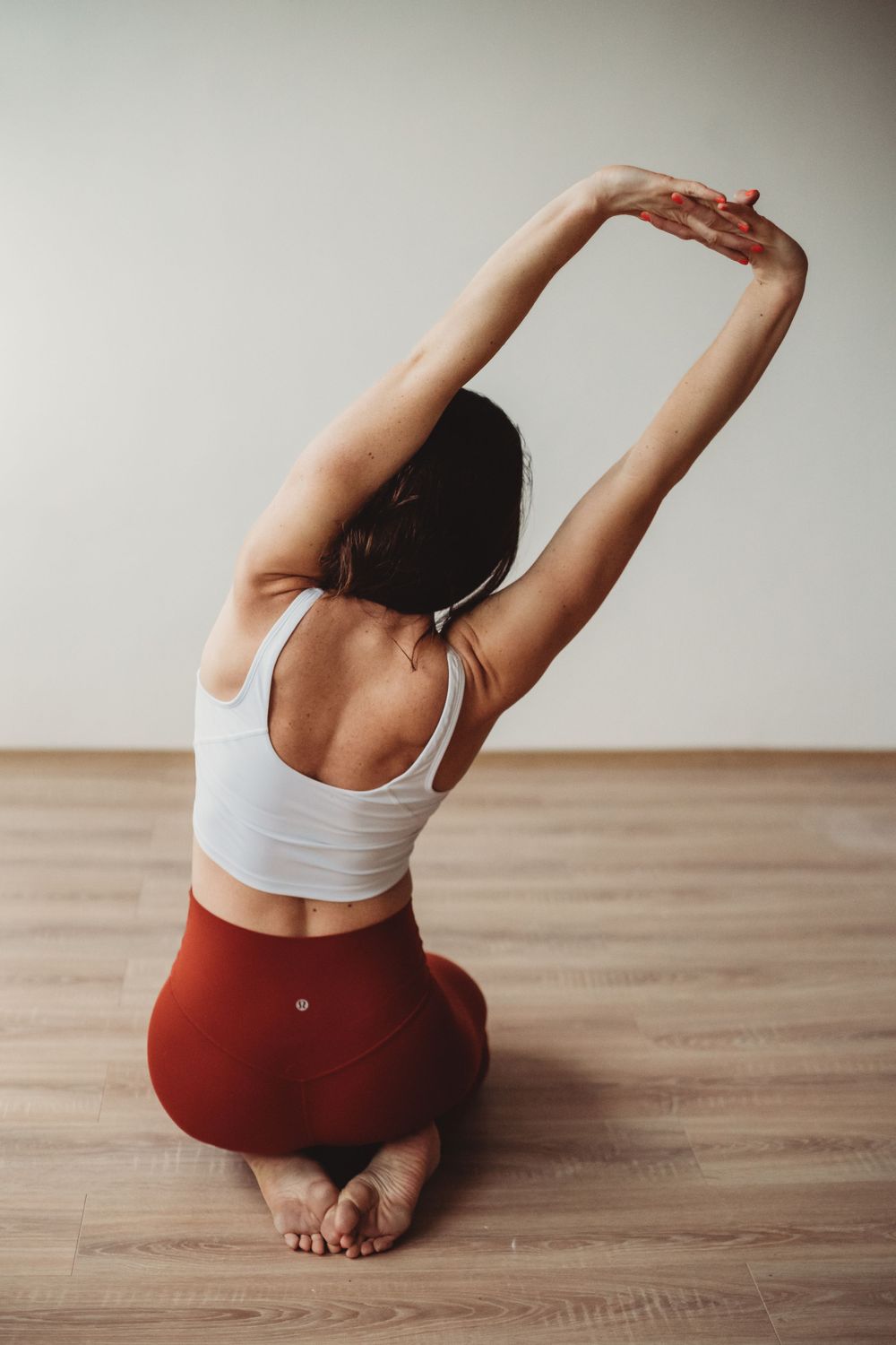 Person in white sports bra and red leggings stretches with arms raised while sitting on wooden floor.