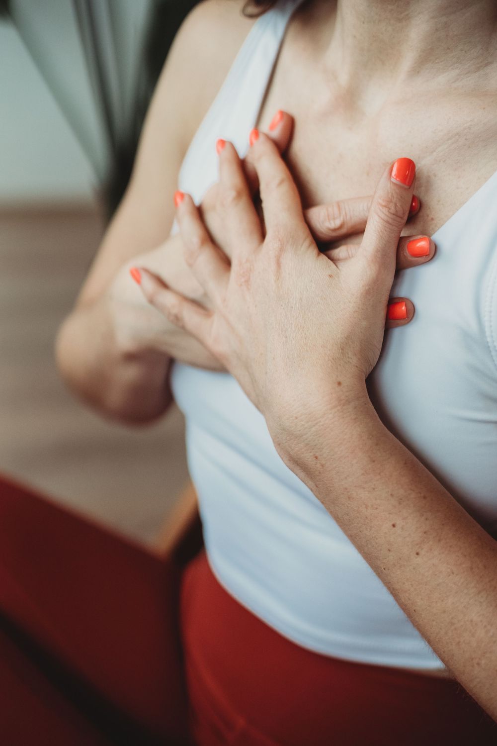 Close-up of hands with bright orange nail polish clutching chest over white tank top in emotional gesture.