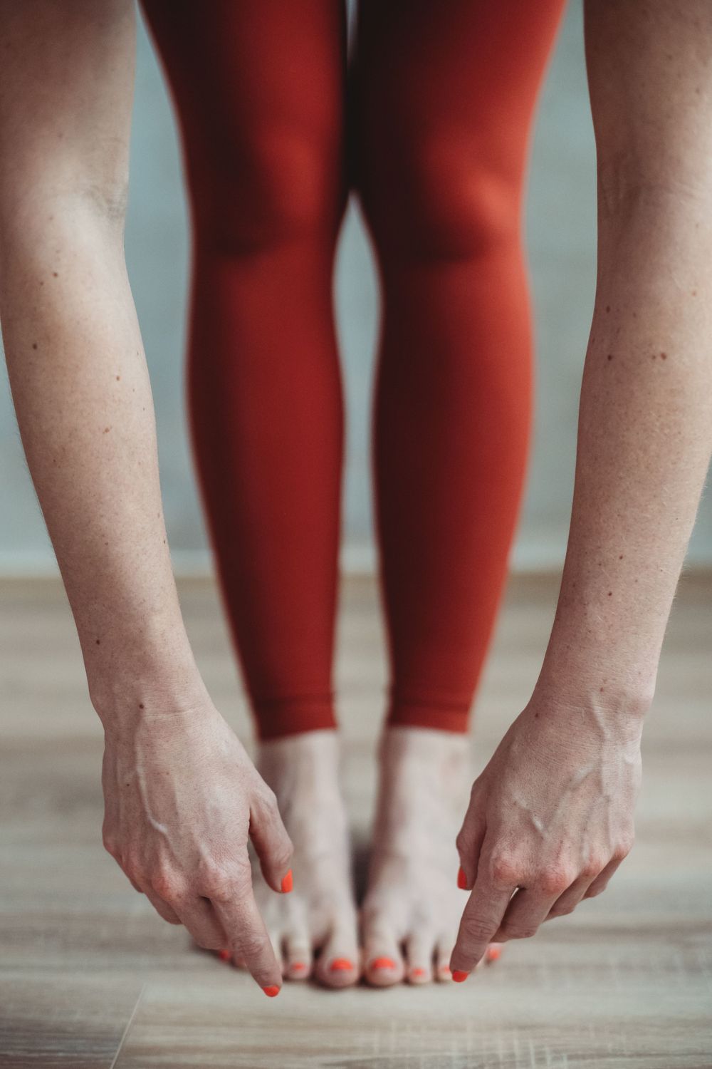 Close-up view of hands reaching down to bare feet while wearing red leggings on a wooden floor.