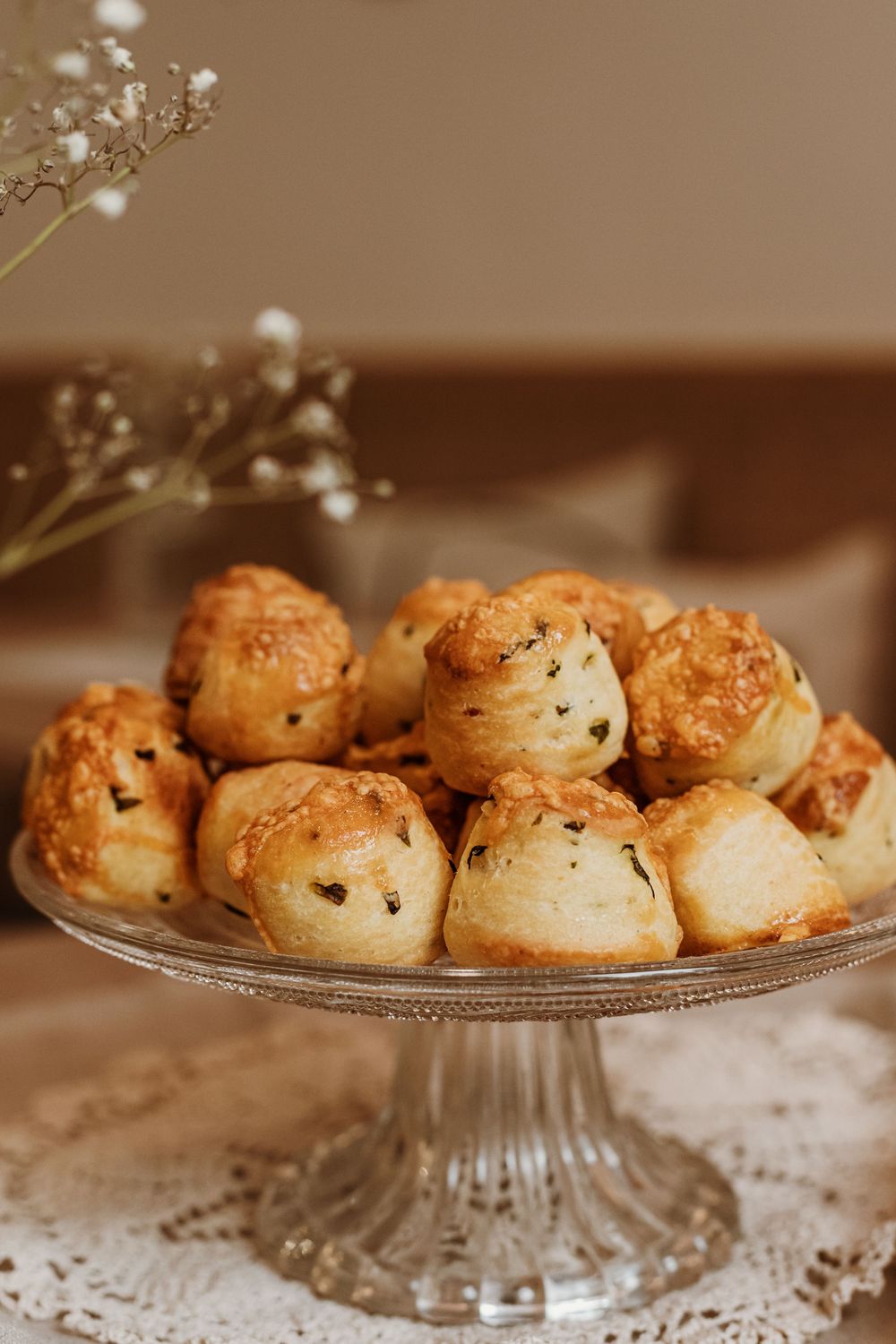 Freshly baked chocolate chip cream puffs displayed on an elegant glass cake stand with baby's breath flowers in background.