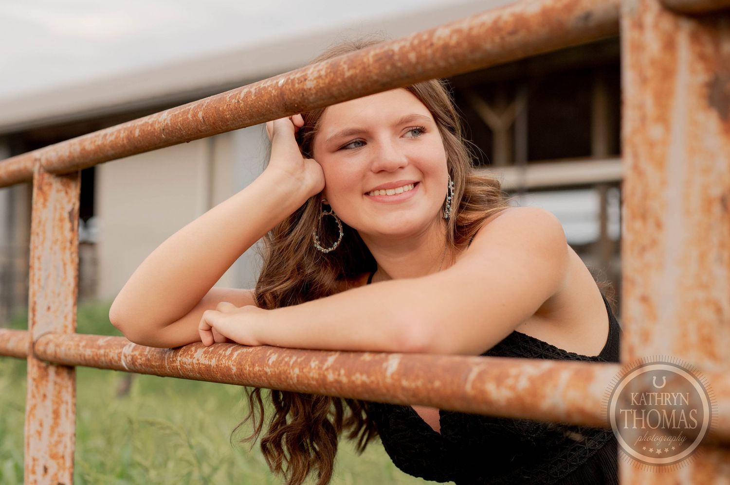 Summer portrait leaning on rustic fence rail with farm buildings behind.
