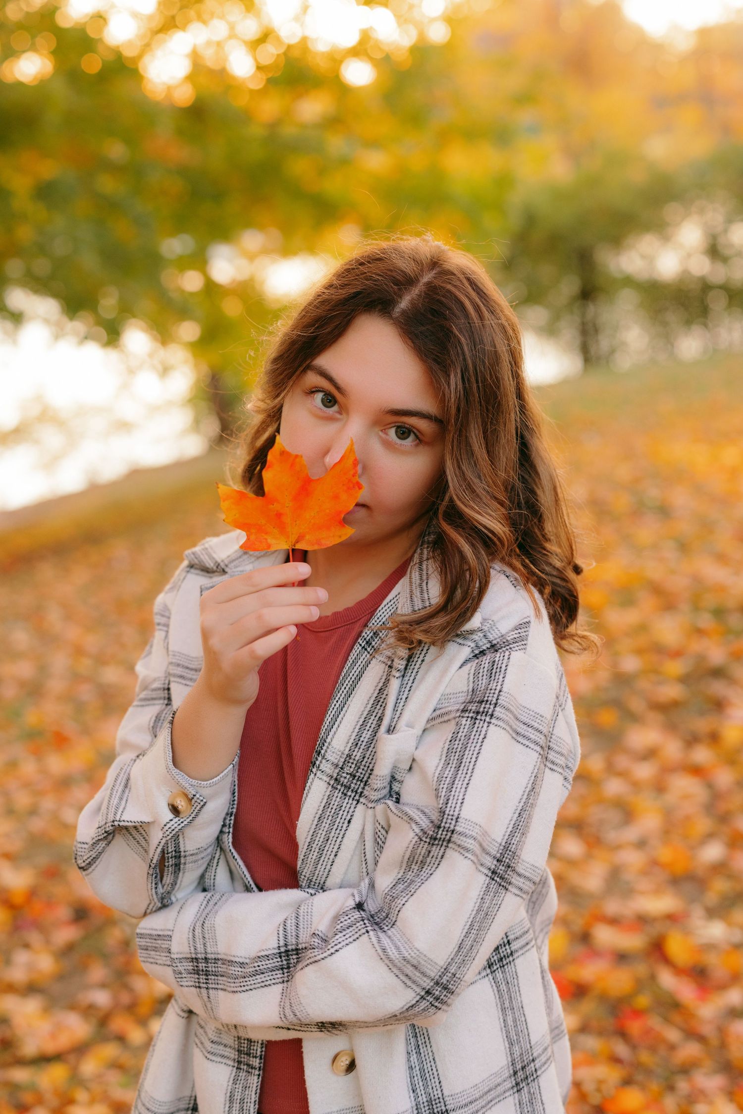 Autumn portrait with a white plaid overshirt and orange maple leaf against golden sunset bokeh in a fall park.
