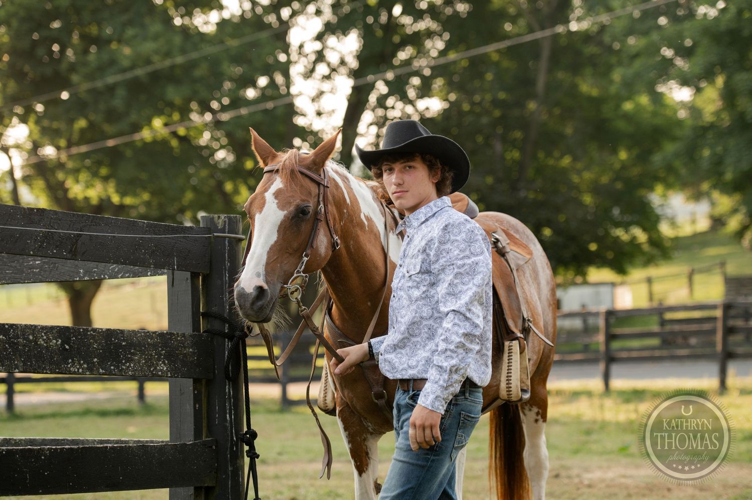 A person in cowboy attire stands with a paint horse near wooden fencing.