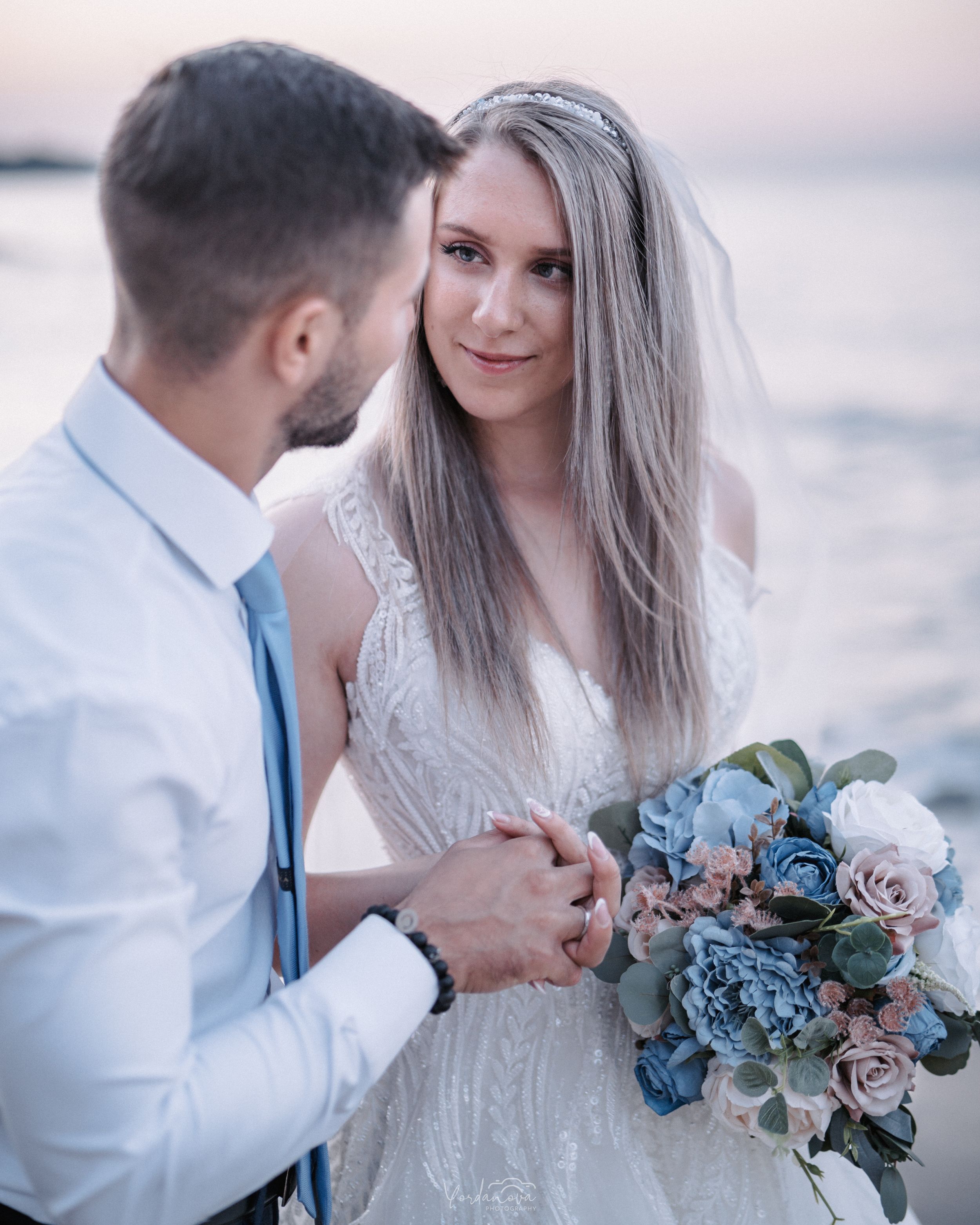 Couple shares intimate moment at sunset while holding floral bouquet by the water.