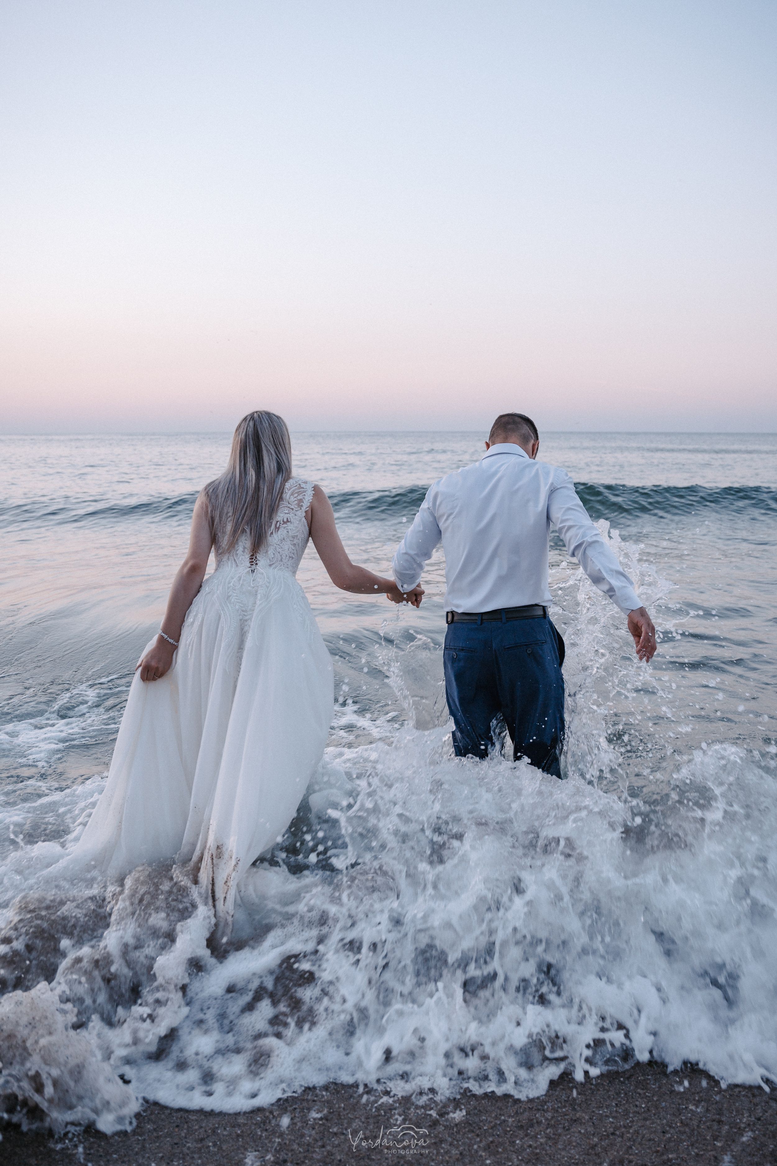 A couple plays in the ocean waves during sunset with flowing white fabric.