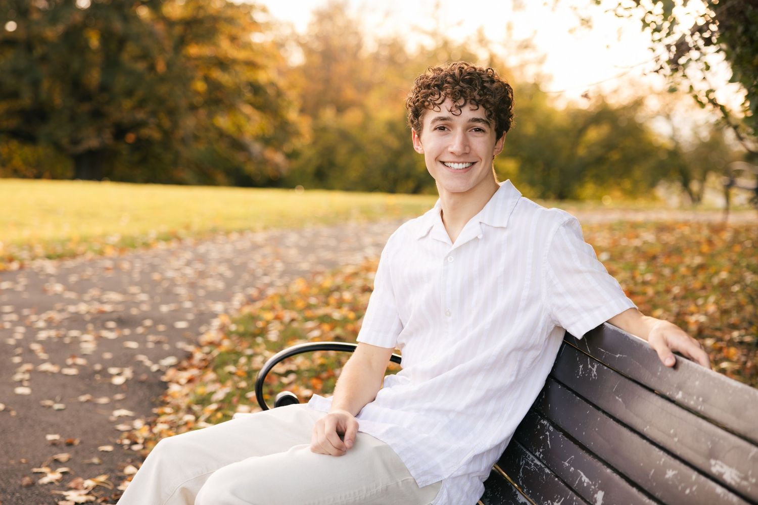 A person wearing a white shirt sits smiling on a wooden park bench during golden hour with autumn foliage in the background.