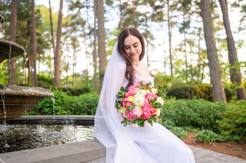 Bride in white gown and veil holding pink bouquet sits near garden fountain surrounded by lush greenery.