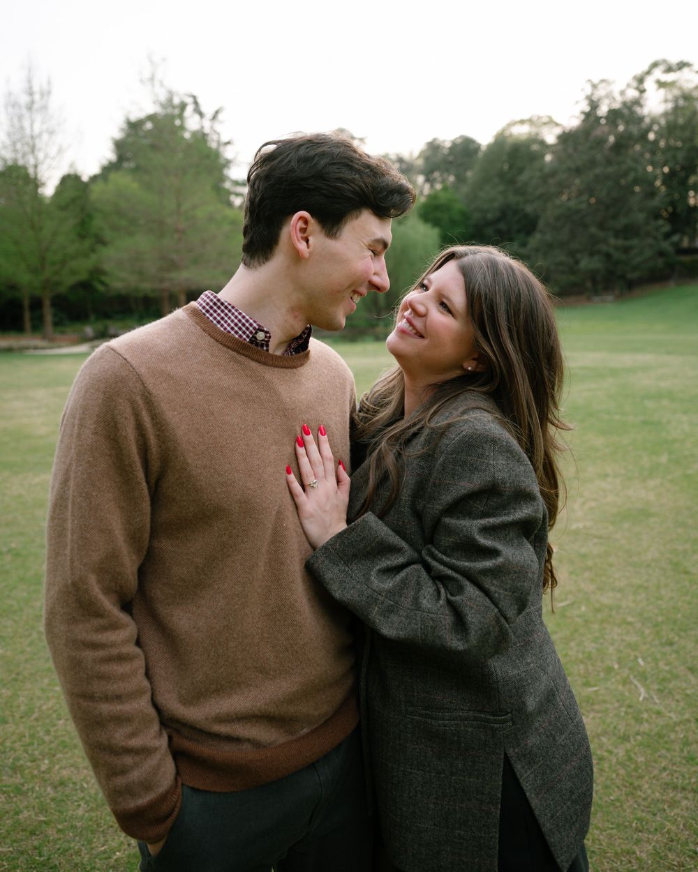 A couple smiles at each other in a lush green park, the woman placing her hand on the man's chest.