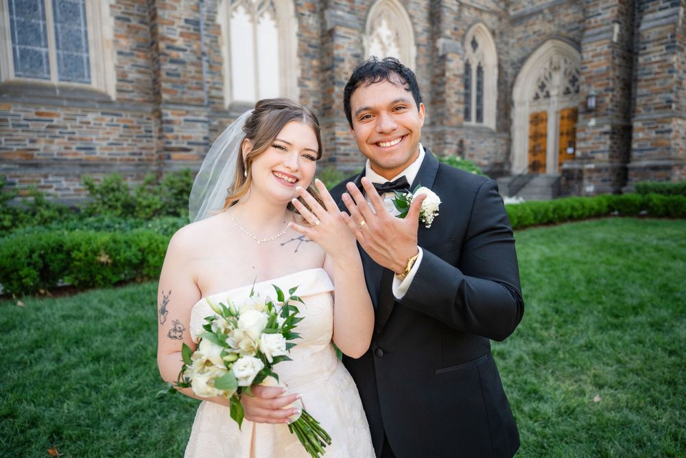 Bride and groom smiling and showing wedding rings outside a stone church, bride holding white bouquet in wedding dress.