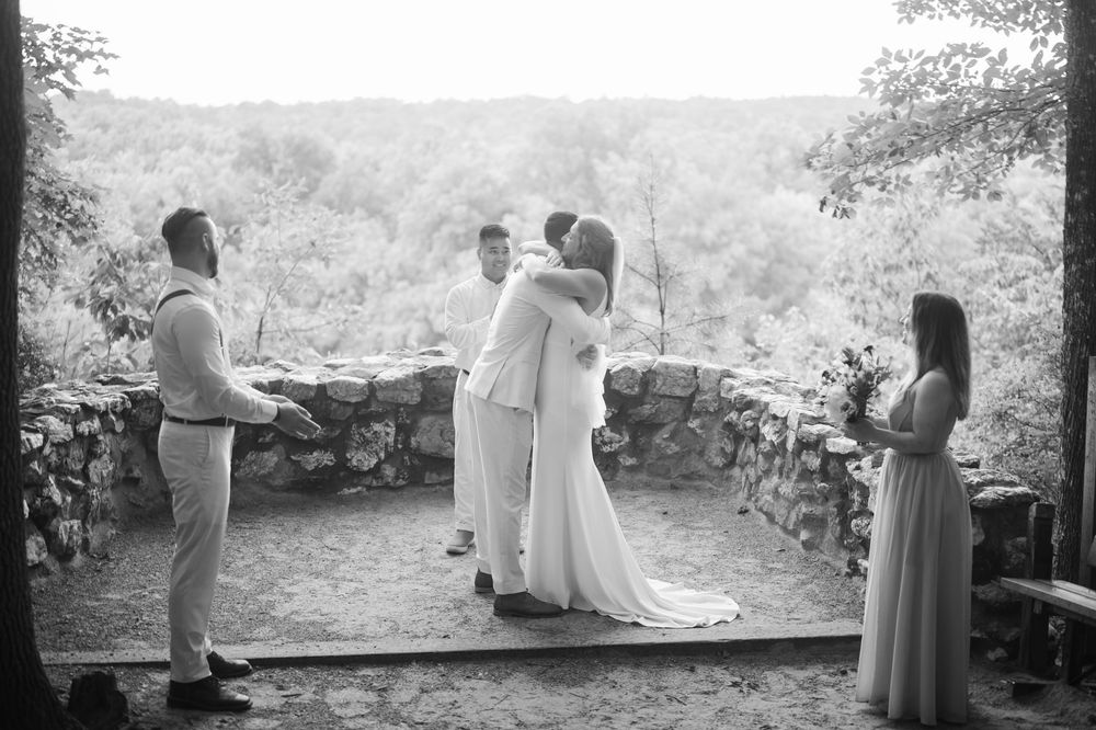 Black and white photo of a bride and groom sharing a first kiss at an outdoor wedding ceremony with stone walls.