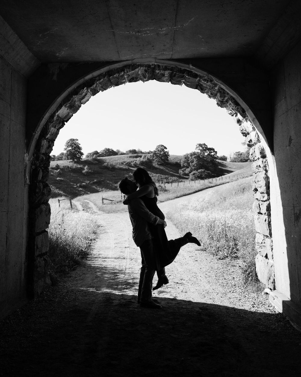 Black and white photo of a couple embracing and kissing silhouetted in a stone arch tunnel with scenic hills beyond.