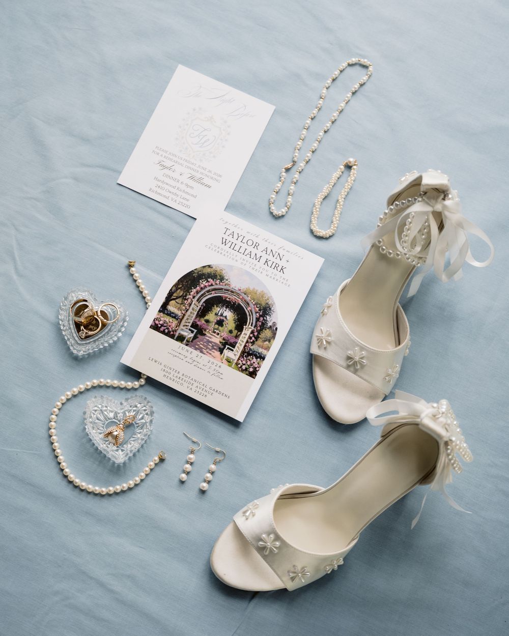 Elegant bridal flat lay featuring white lace heels, pearl jewelry, gold rings, and a wedding program on a soft blue background.