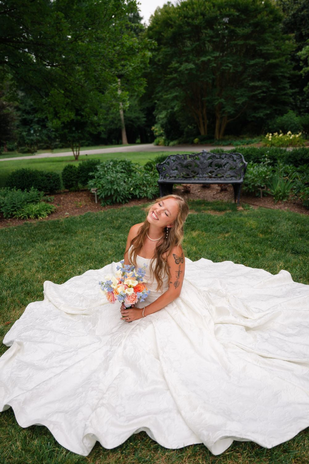 Bride in a voluminous white ball gown sits on green grass, laughing joyfully while holding a colorful bouquet in a garden setting.