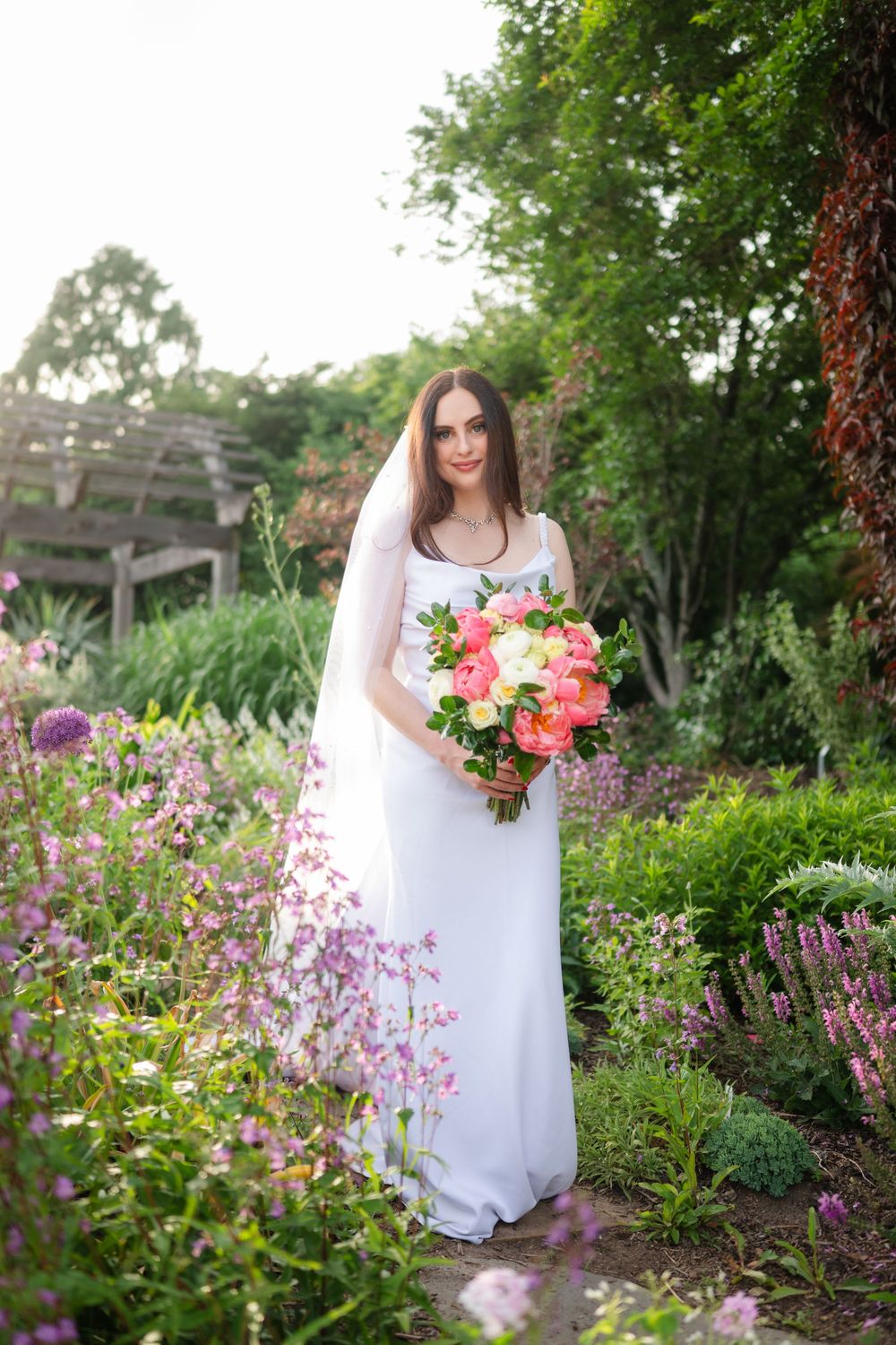 Bride in white gown and veil holds colorful bouquet in lush garden with purple and pink flowers.