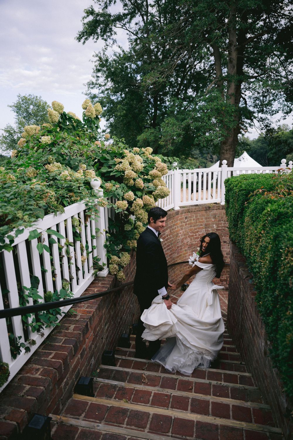 Bride and groom walking down brick stairs surrounded by white hydrangeas and lush greenery at an outdoor wedding venue.