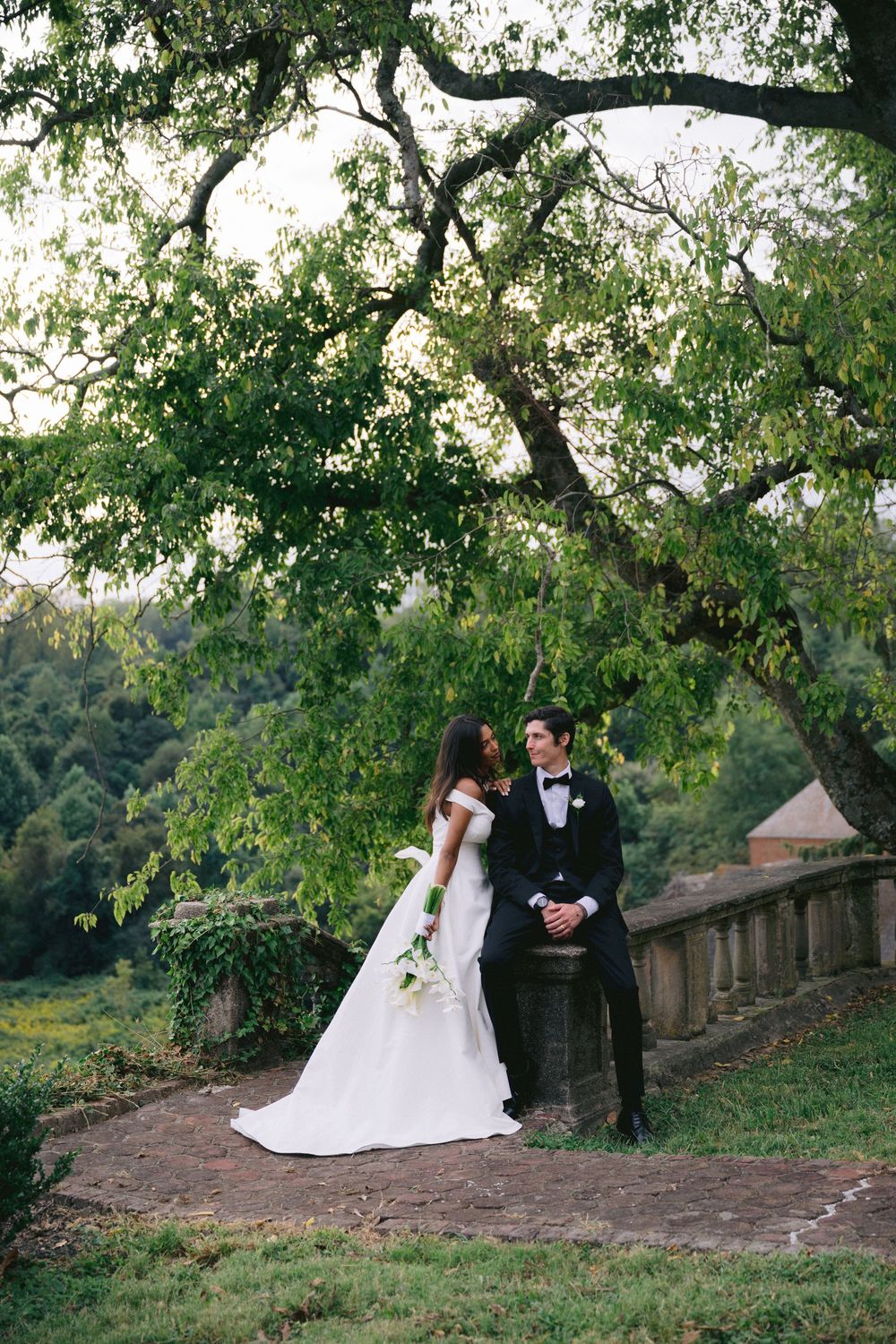 Bride in white gown and groom in black tuxedo pose under lush green trees on a stone terrace at their outdoor wedding.