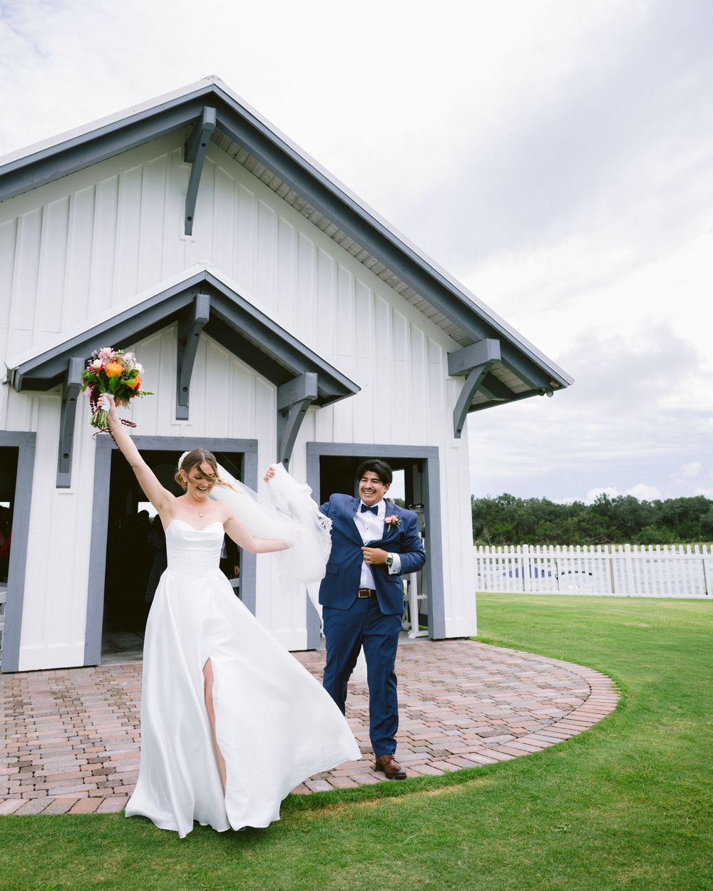Bride raises bouquet while groom in navy suit celebrates outside white barn wedding venue on lush green lawn.