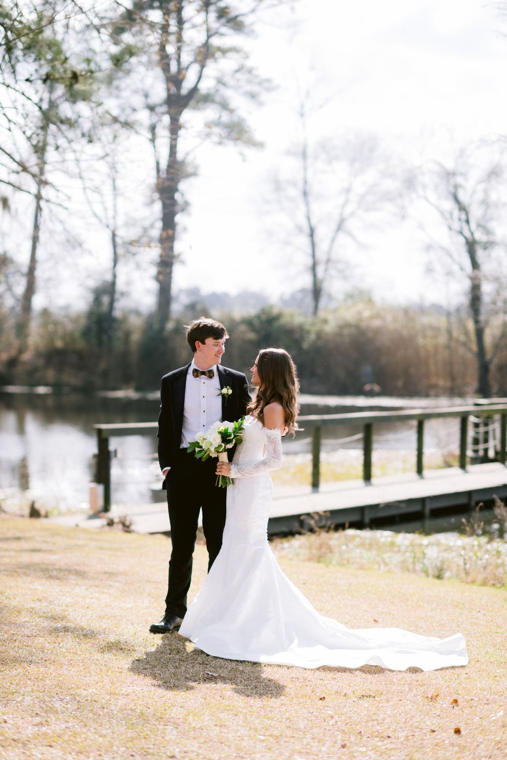 Bride and groom share a romantic moment by a lakeside dock, she in a white gown, he in a black tuxedo.