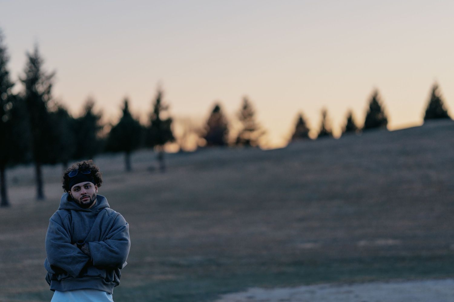 A person in winter clothing stands on a hillside against evergreen trees at dusk.