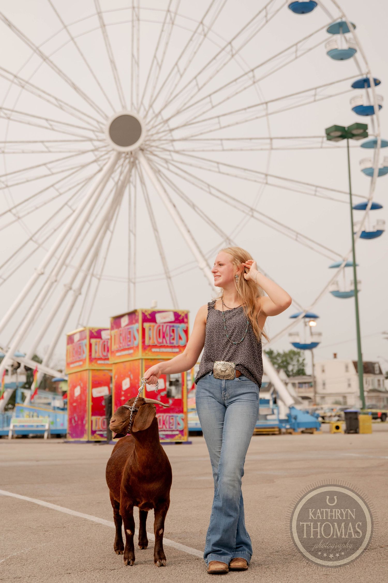 Goat and handler walk past ferris wheel at sunset during county fair.