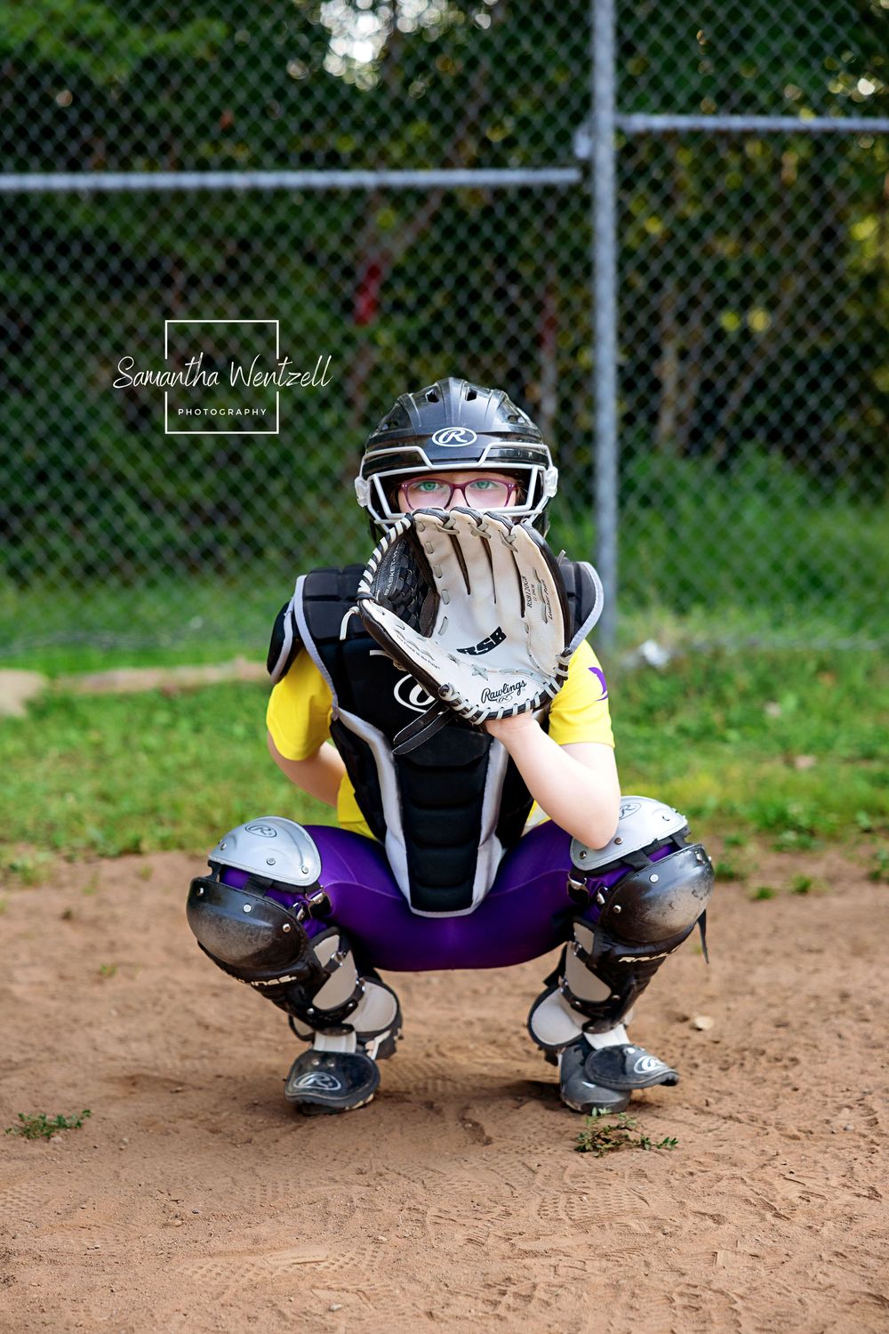 Baseball catcher in protective gear crouches low to the ground in ready position on a dirt field.