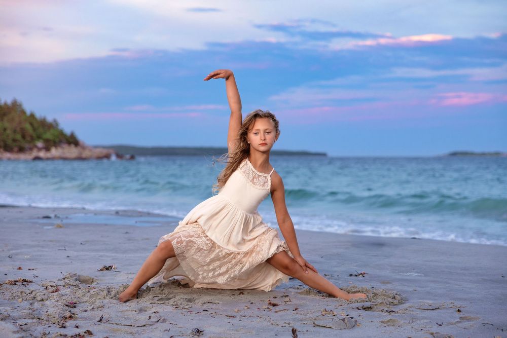 A dancer in a flowing white dress performs an elegant pose on a beach at sunset with turquoise waters and cliffs in background.