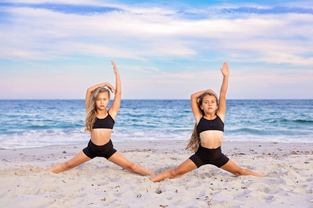 Two dancers in black athletic wear perform synchronized splits poses on a sunny beach with ocean waves in background.