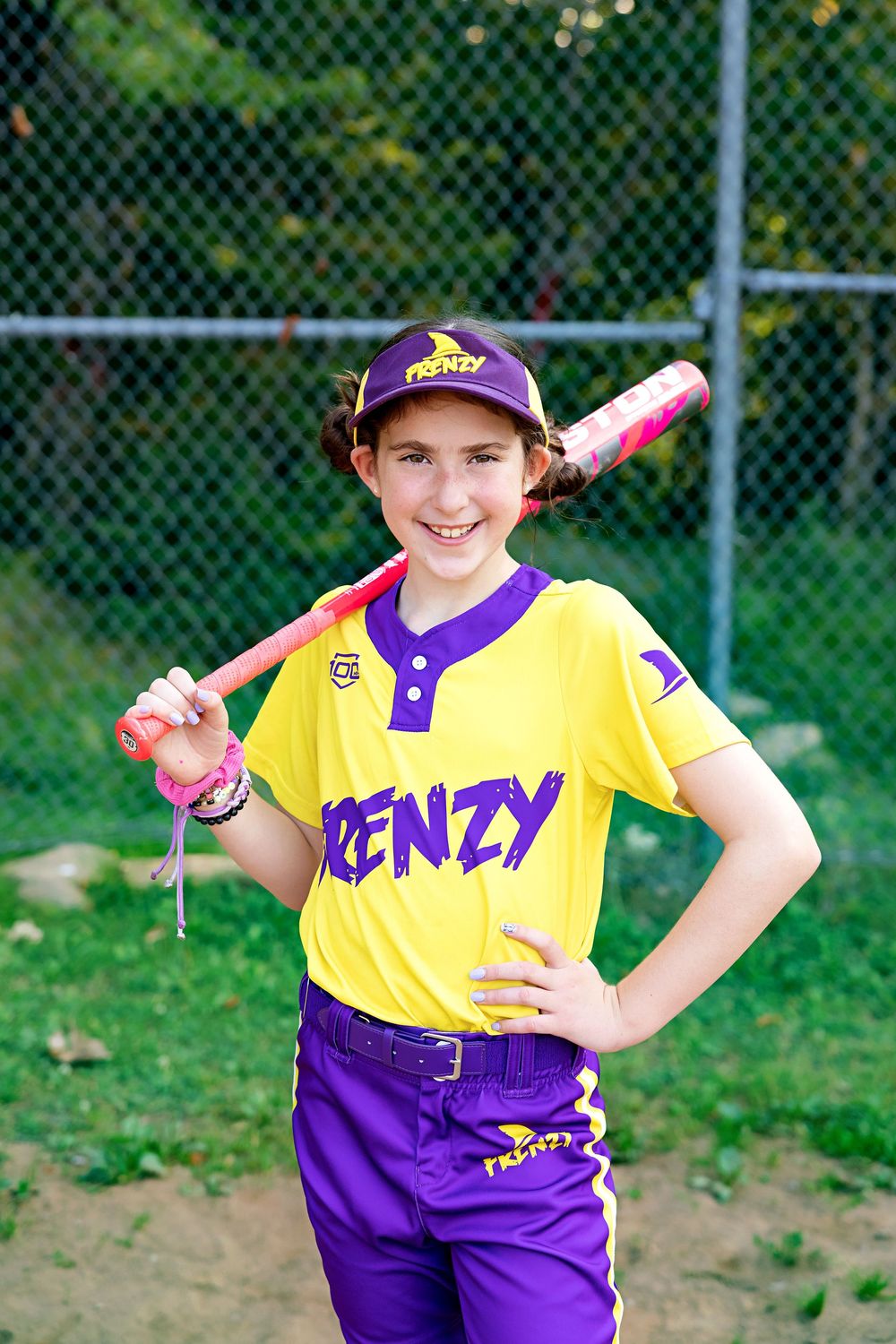 Young softball player in yellow and purple uniform poses with a bat on a sunny day at the baseball field.