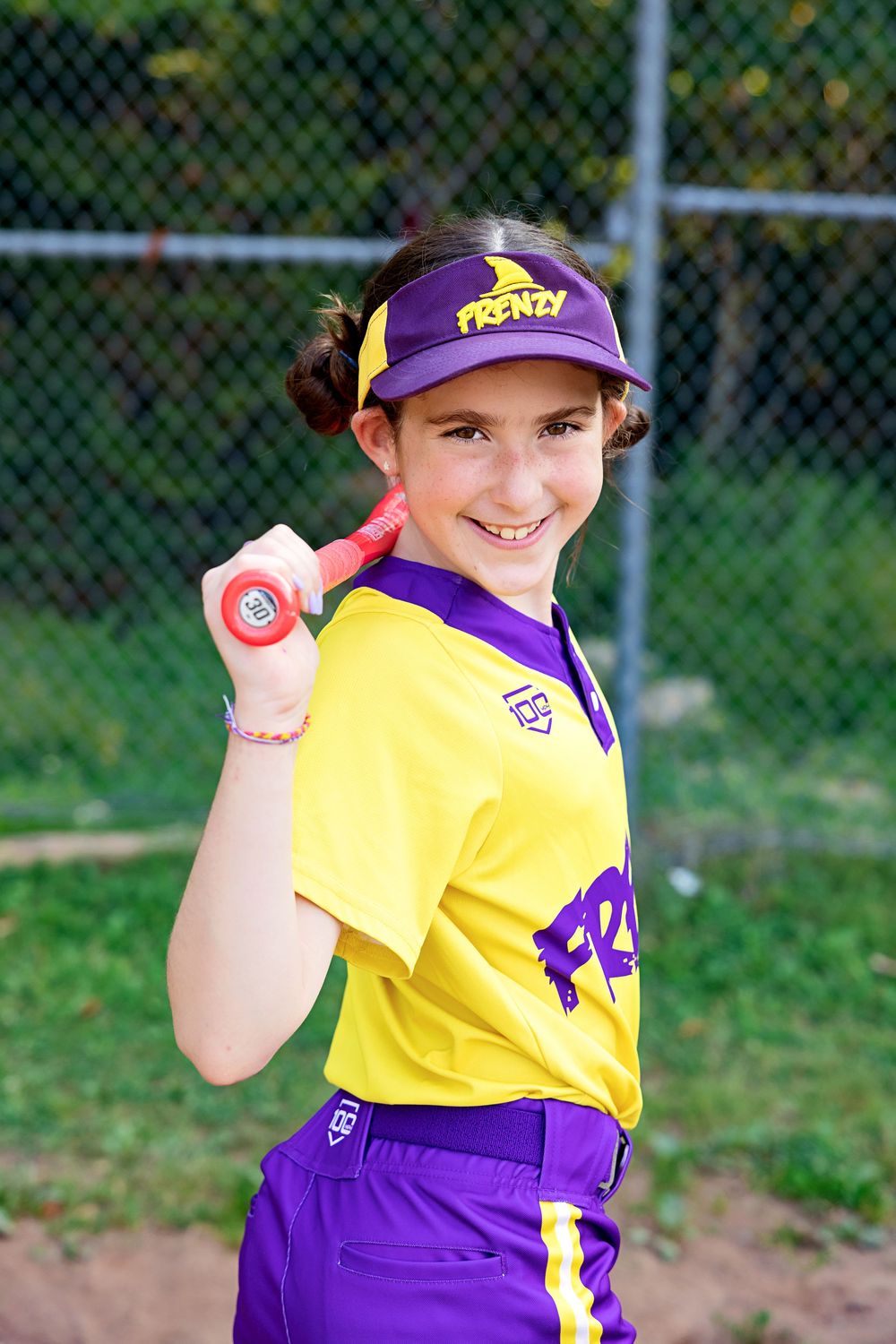 Young softball player in purple and yellow uniform shows baseball stance while practicing outdoors.